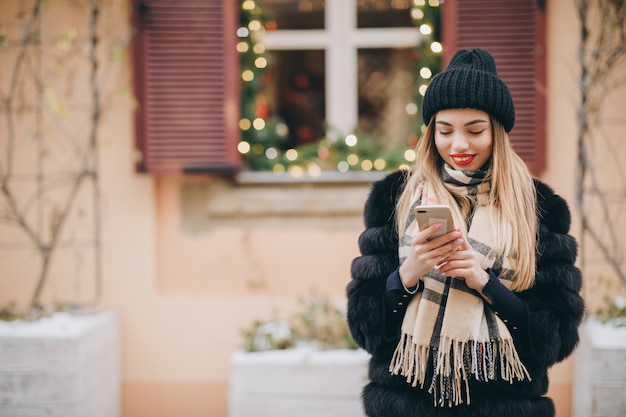 Ritratto di giovane donna felice che indossa abiti invernali in stile francese, utilizzando smartphone