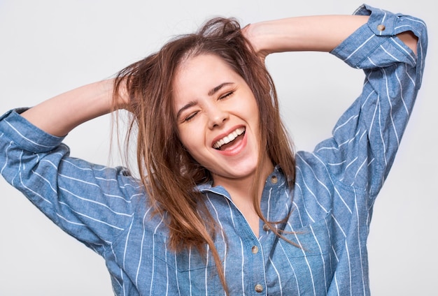 Ritratto di giovane donna felice che indossa una camicia blu a righe casual sorridente ampiamente con la bocca spalancata Ragazza positiva che si diverte con le mani sui capelli in posa in studio Emozioni della gente