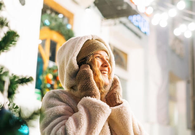 Ritratto di giovane donna felice con i capelli ricci in giacca di pelliccia divertendosi nella strada invernale decorata con luci