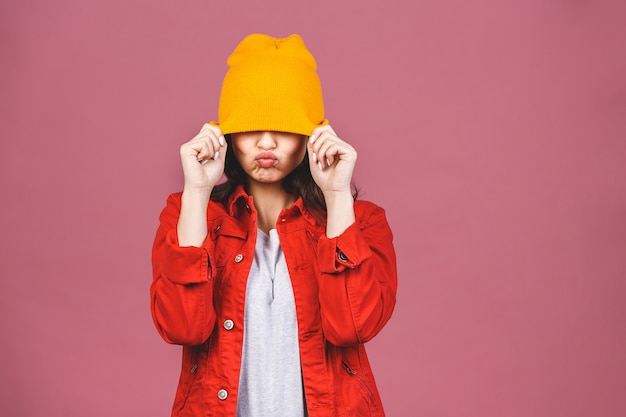 Ritratto di giovane donna felice dei pantaloni a vita bassa in cappello giallo e camicia rossa isolati sopra la parete rosa.