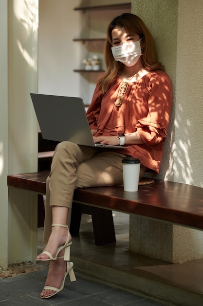 Ritratto di giovane donna felice in maschera protettiva seduta su una panchina con una tazza di caffè da asporto e codifica sul computer portatile