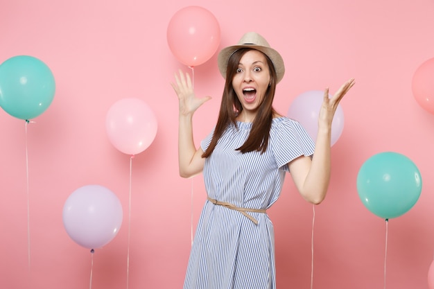 Ritratto di giovane donna felice sorpresa che indossa cappello estivo di paglia e vestito blu che allarga le mani su sfondo rosa pastello con mongolfiere colorate. Festa di compleanno persone emozioni sincere.