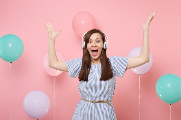 Ritratto di giovane donna felicissima con la bocca aperta con le cuffie che indossano abiti blu ascoltando musica diffondendo le mani su sfondo rosa con mongolfiere colorate. Concetto di festa di compleanno.