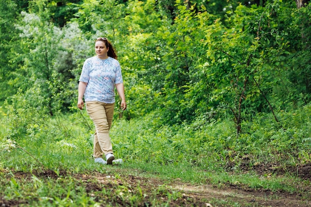 Ritratto di giovane donna grassa con lunghi capelli rossi ricci che cammina sul sentiero nella foresta del parco tra alberi verdi Fine settimana estivo