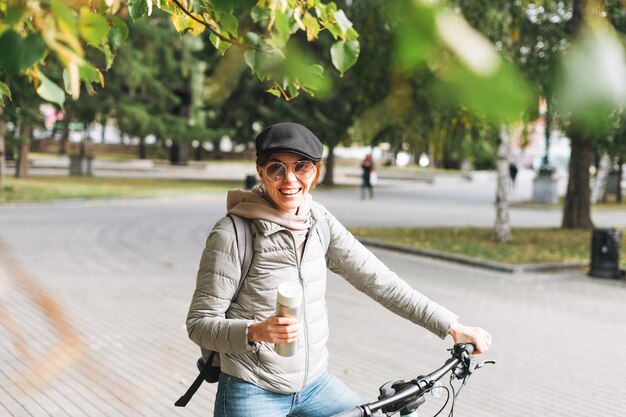 Ritratto di giovane donna graziosa alla moda in berretto e occhiali da sole in bicicletta con tazza termica in una soleggiata giornata autunnale nel parco cittadino