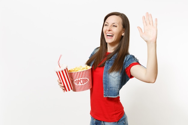 Ritratto di giovane donna graziosa che ride in abiti casual guardando film, tenendo secchio di popcorn, tazza di plastica di soda o cola che mostra palmo in su isolato su priorità bassa bianca. Emozioni al cinema.