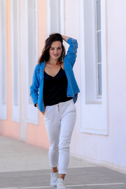 Ritratto di giovane donna graziosa sorridente allegra sveglia in vestiti casuali dei jeans. In piedi sullo sfondo del corridoio chiaro.