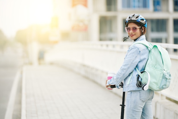 Ritratto di giovane donna graziosa sorridente che indossa un casco protettivo quando si guida su uno scooter elettrico con un grande zaino