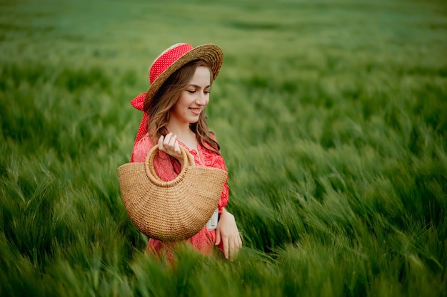 Ritratto di giovane donna in abito e cappello nel campo verde di orzo in campagna. Ragazza alla moda nel momento pacifico godente rustico in erba di estate.