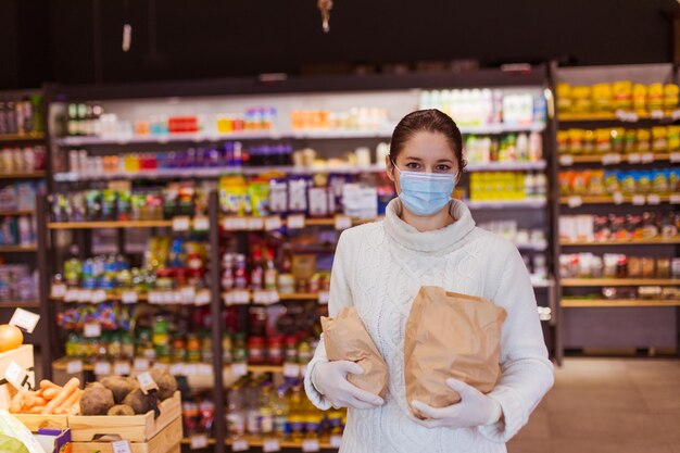 Ritratto di giovane donna in caldo sudore che indossa maschera protettiva e guanti con in mano due sacchetti di carta con cibo Scaffali con vari prodotti sullo sfondo Sostenere il concetto di business locale