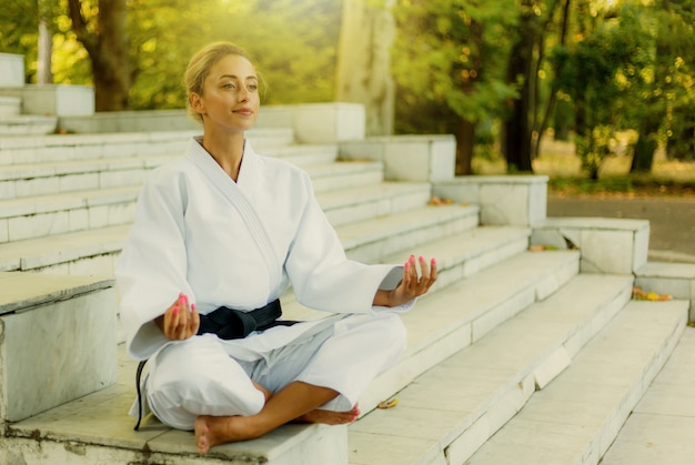 Ritratto di giovane donna in kimono bianco con cintura nera. Sport donna seduta sulle scale e meditare all'aperto. Arti marziali