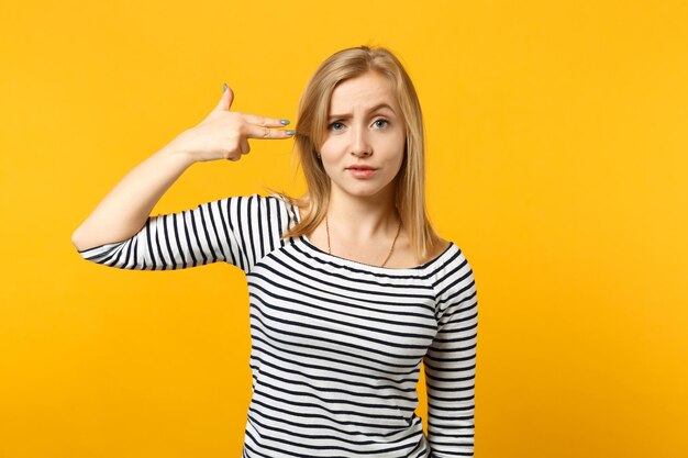 Ritratto di giovane donna insoddisfatta in abiti a righe punta le dita verso la testa come se stesse per spararsi isolata su sfondo giallo arancio in studio. Persone sincere emozioni, concetto di stile di vita.