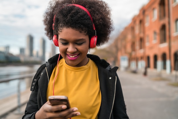 Ritratto di giovane donna latina afroamericana che utilizza telefono cellulare e che ascolta la musica con le cuffie nella via. All'aperto.