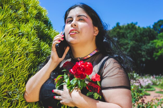 Ritratto di giovane donna latina ispanica caucasica innamorata drammatica in un parco pubblico con rose e alberi che sta parlando al telefono abbracciando alcuni fiori