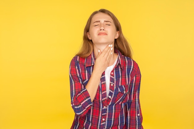 Ritratto di giovane donna malata in camicia a scacchi che tocca il collo dolorante, fa smorfie per il dolore, tonsille doloranti, disturbi della tiroide e sintomi influenzali. tiro in studio indoor isolato su sfondo giallo