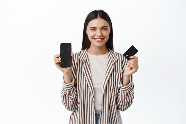 Ritratto di giovane donna moderna in abito elegante, che mostra lo schermo vuoto dello smartphone e una carta di credito in plastica, sorridendo soddisfatto, consigliando un'app, muro bianco