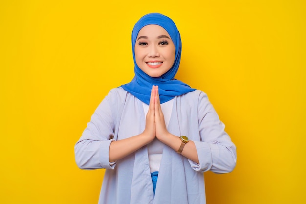 Ritratto di giovane donna musulmana asiatica sorridente che gesturing il saluto di Eid Mubarak isolato su sfondo giallo