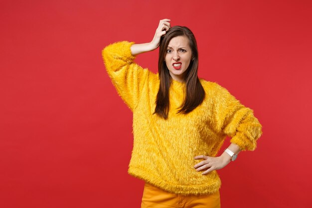 Ritratto di giovane donna perplessa interessata in maglione di pelliccia gialla che mette mano sulla testa isolata su fondo rosso brillante della parete in studio. Persone sincere emozioni, concetto di stile di vita. Mock up copia spazio.