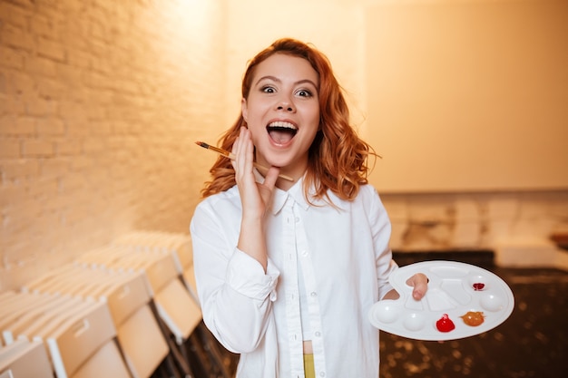 Ritratto di giovane donna pittrice rossa felice scioccata con colori ad olio e tavolozza.