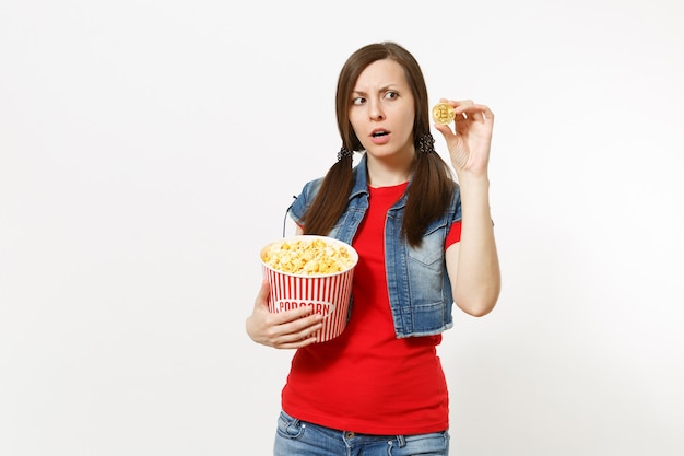 Ritratto di giovane donna piuttosto bruna interessata in abiti casual che guarda film, tenendo in mano un secchio di popcorn e bitcoin dorato isolato su sfondo bianco. Emozioni nel concetto di cinema.