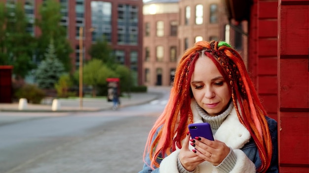 Ritratto di giovane donna positiva con i dreadlocks che naviga sul cellulare in piedi appoggiato al muro sulla strada della città