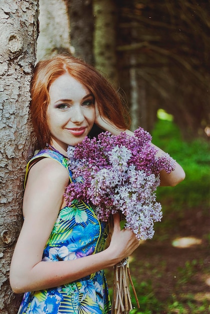 Ritratto di giovane donna rossa nel giardino di primavera. lentiggini. fiori viola in fiore. Mazzo di lillà