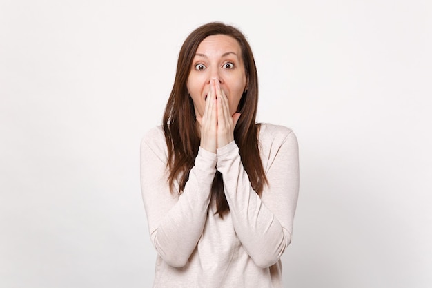 Ritratto di giovane donna scioccata in abiti leggeri che guarda la fotocamera, coprendo la bocca con le mani isolate su sfondo bianco della parete in studio. Persone sincere emozioni, concetto di stile di vita. Mock up spazio di copia.