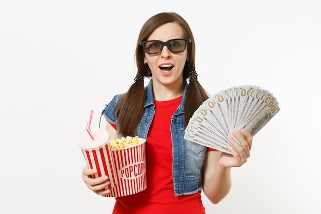 Ritratto di giovane donna scioccata in occhiali 3d guardando film, tenendo secchio di popcorn, tazza di soda o cola e pacco di dollari, denaro contante isolato su sfondo bianco. Emozioni al cinema.