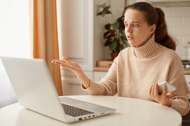 Ritratto di giovane donna seria adulta che indossa un maglione beige in posa in cucina e lavora al computer portatile con un'importante conversazione online che spiega i dettagli del progetto per il suo partner