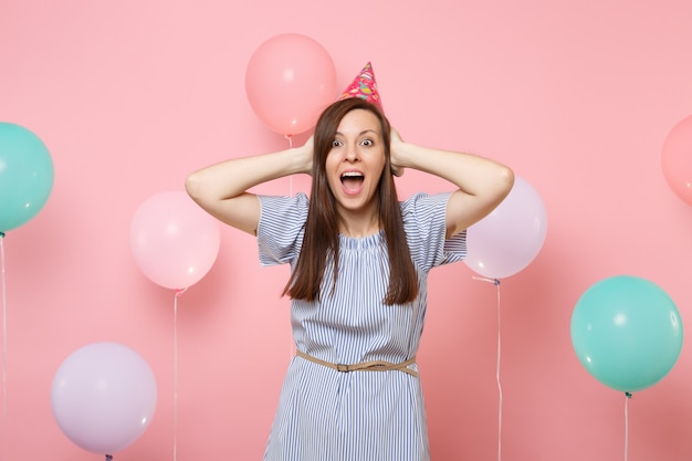 Ritratto di giovane donna sorpresa in cappello di compleanno e vestito blu che screming coprendo le orecchie su sfondo rosa pastello con palloncini colorati. Festa di compleanno, concetto di emozioni sincere della gente.