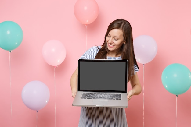 Ritratto di giovane donna sorridente attraente che indossa vestito blu che tiene il computer portatile del pc con lo schermo vuoto in bianco su fondo rosa pastello con gli aerostati di aria variopinti. Concetto di festa di compleanno.