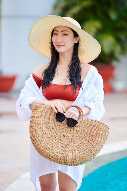 Ritratto di giovane donna sorridente attraente in cappello di paglia, bikini e camicia di cotone bianca in piedi in piscina con borsa di vimini e occhiali da sole nelle mani