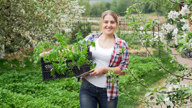 Ritratto di giovane donna sorridente con cassa piena di piantine in posa nel giardino del cortile.