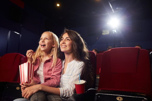 Ritratto di giovane donna sorridente con figlia carina guardando cartoni animati al cinema mentre guarda lo schermo e godendo di popcorn, copia spazio