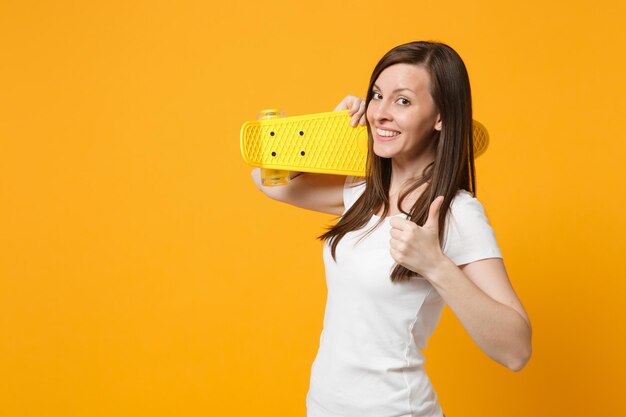 Ritratto di giovane donna sorridente in abiti casual bianchi che mostra il pollice in su, tenendo lo skateboard giallo isolato su sfondo giallo arancione brillante in studio. Concetto di stile di vita delle persone. Mock up spazio di copia.