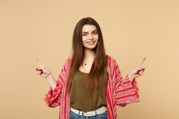 Ritratto di giovane donna sorridente in abiti casual che guarda la macchina fotografica, puntando il dito indice da parte isolato sul fondo della parete beige pastello. Persone sincere emozioni, concetto di stile di vita. Mock up copia spazio.