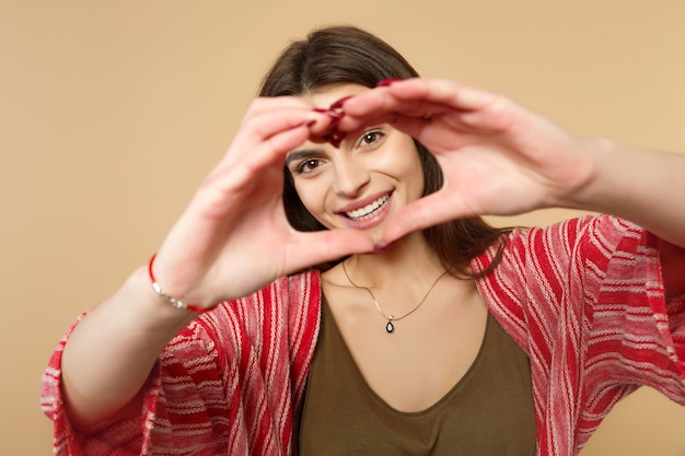 Ritratto di giovane donna sorridente in abiti casual che mostra il cuore di forma con le mani isolate sul fondo della parete beige pastello in studio. Persone sincere emozioni, concetto di stile di vita. Mock up copia spazio.