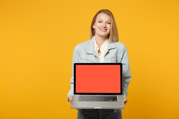 Ritratto di giovane donna sorridente in abiti casual denim in possesso di computer portatile pc con schermo vuoto vuoto isolato su sfondo giallo arancio in studio. Concetto di stile di vita delle persone. Mock up spazio di copia.