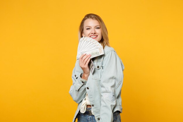 Ritratto di giovane donna sorridente in abiti casual denim tenere fan di denaro contante in banconote in dollari isolate su sfondo giallo muro arancione in studio. Concetto di stile di vita delle persone. Mock up spazio di copia.