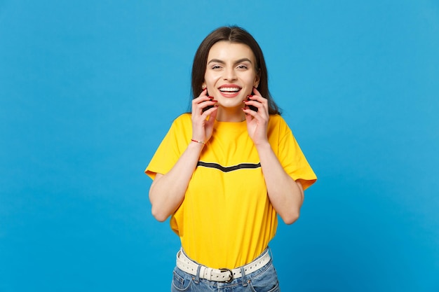 Ritratto di giovane donna sorridente in abiti casual vivaci che guarda la fotocamera, mettendo le mani vicino al viso isolato su uno sfondo blu brillante della parete in studio. Concetto di stile di vita delle persone. Mock up spazio di copia.