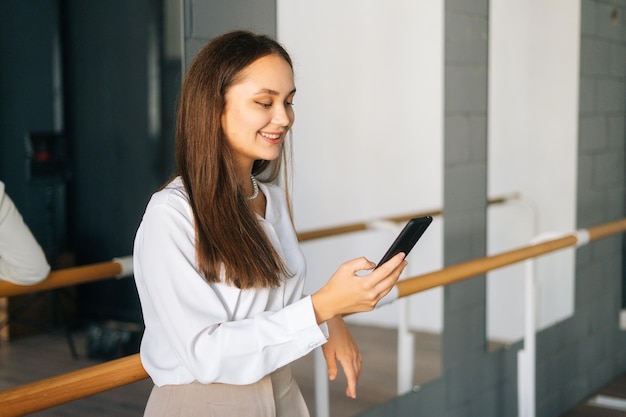 Ritratto di giovane donna sorridente in abiti eleganti utilizzando il telefono cellulare di navigazione in piedi nella stanza con specchio che guarda sullo schermo