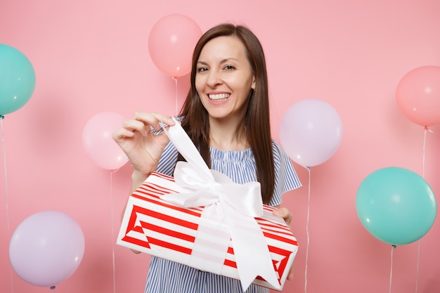 Ritratto di giovane donna sorridente in abito blu che slega l'arco sulla scatola rossa con regalo presente su sfondo rosa pastello con mongolfiere colorate. Festa di compleanno, concetto di emozioni sincere della gente.