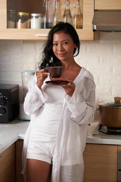 Ritratto di giovane donna sorridente in loungewear in piedi in cucina e bere una tazza di caffè mattutino