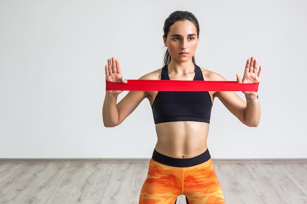 Ritratto di giovane donna sportiva che indossa top nero e leggings arancioni esegue esercizi per i muscoli delle mani, allenamento con fascia di resistenza su sfondo bianco. Forza e motivazione. Interno