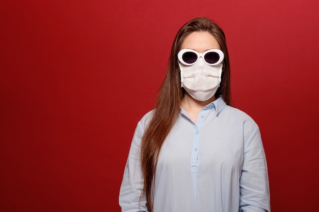 Ritratto di giovane donna sulla parete rossa in maschera protettiva medica e occhiali da sole