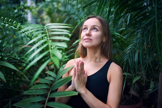 Ritratto di giovane donna sveglia che medita e che fa la mano del namaste in giungla.