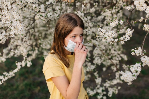 Ritratto di giovane donna triste in maschera protettiva medica con fiori vicino albero fiorito in primavera. Concetto di allergia di primavera