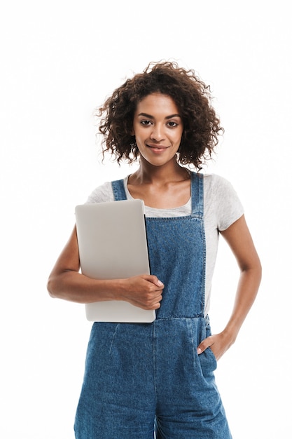 Ritratto di giovane donna vestita con una tuta di jeans che sorride e tiene in mano un laptop isolato su un muro bianco