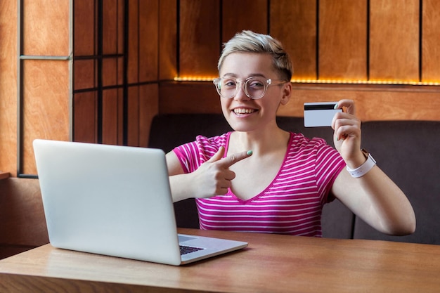 Ritratto di giovane e bella bussineswoman felice con i capelli corti in maglietta rosa e occhiali da vista è seduto al bar, puntando il dito sulla carta di credito, facile da ordinare online, guardando la fotocamera. Interno