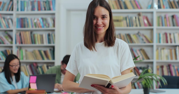 Ritratto di giovane e bella ragazza in possesso di un libro sullo sfondo dello scaffale della biblioteca guardando la fotocamera Concetto di istruzione universitaria